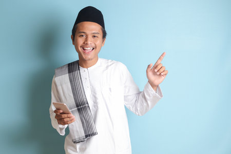 Portrait of young Asian muslim man holding mobile phone with smiling expression on face while pointing finger to the side. Isolated image on blue backgroundの写真素材