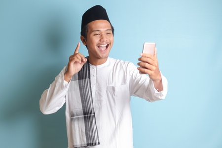 Portrait of Asian muslim man in white koko shirt with skullcap taking picture of himself or selfie, saying hi and waving his hand during video call. Isolated image on blue backgroundの写真素材