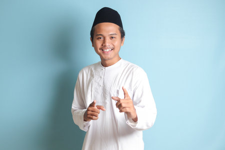 Portrait of young Asian muslim man showing product and pointing with his hand and finger to the side. Isolated image on blue backgroundの写真素材