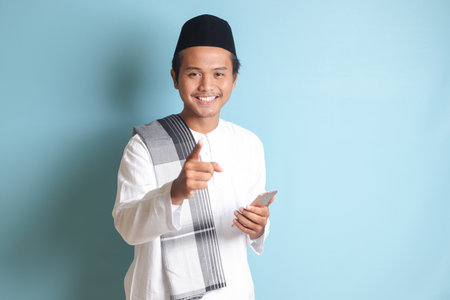 Portrait of young Asian muslim man holding mobile phone with smiling expression on face while pointing finger to the side. Isolated image on blue backgroundの写真素材