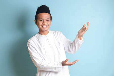 Portrait of young Asian muslim man showing product and pointing with his hand and finger to the side. Isolated image on blue backgroundの写真素材