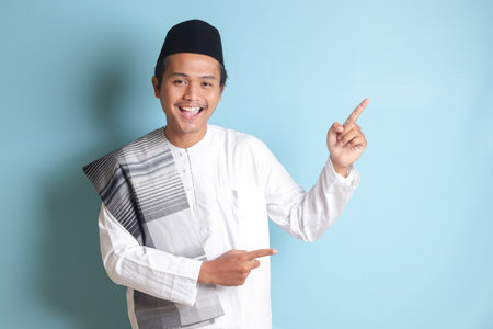 Portrait of young Asian muslim man showing product and pointing with his hand and finger to the side. Isolated image on blue backgroundの写真素材