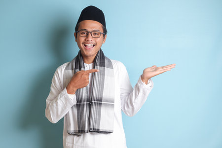 Portrait of young Asian muslim man showing product and pointing with his hand and finger to the side. Isolated image on blue backgroundの写真素材