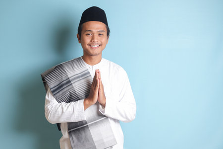 Portrait of Asian muslim man in white koko shirt with skullcap showing apologize and welcome hand gesture. Isolated image on blue backgroundの写真素材