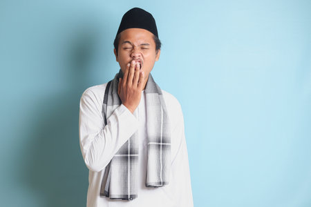 Portrait of attractive Asian muslim man in white shirt yawning with hand covering his mouth. Isolated image on blue backgroundの写真素材