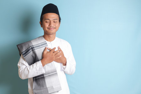 Portrait of religious Asian man in koko shirt or white muslim shirt and black cap, placing hand on heart, feeling very grateful. Isolated image on gray backgroundの写真素材