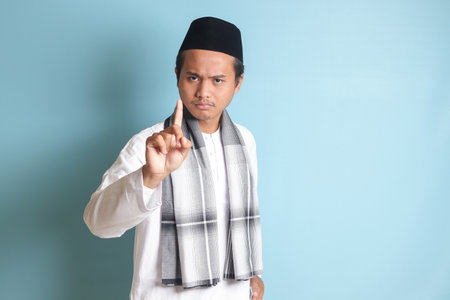 Portrait of young Asian muslim man pointing up his forefinger and reminding something to do. Advertising concept. Isolated image on blue backgroundの写真素材