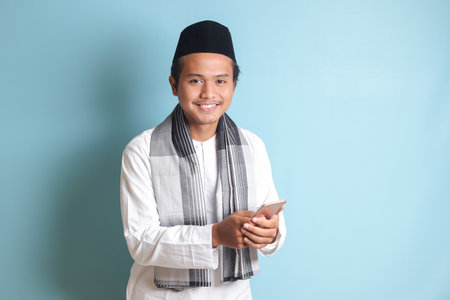 Portrait of young Asian muslim man holding and touching mobile phone with smiling expression on face. Isolated image on blue backgroundの写真素材