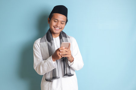 Portrait of young Asian muslim man holding and touching mobile phone with smiling expression on face. Isolated image on blue backgroundの写真素材
