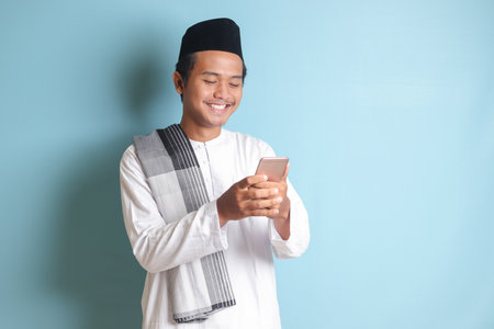 Portrait of young Asian muslim man holding and touching mobile phone with smiling expression on face. Isolated image on blue backgroundの写真素材
