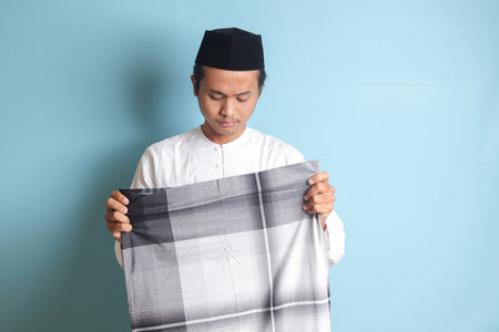 Portrait of Asian muslim man in white koko shirt folding and tidy up his sarung . Isolated image on blue backgroundの写真素材