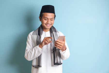 Portrait of young Asian muslim man holding and touching mobile phone with smiling expression on face. Isolated image on blue backgroundの写真素材