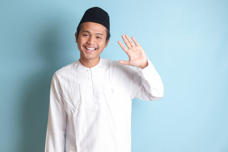 Portrait of Asian muslim man in white koko shirt with skullcap shirt saying hi, waving hand at camera, greeting person with happy smile. Isolated image on blue backgroundの写真素材