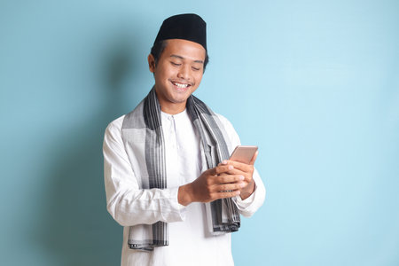 Portrait of young Asian muslim man holding and touching mobile phone with smiling expression on face. Isolated image on blue backgroundの写真素材