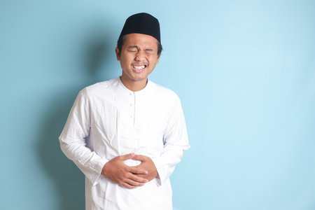 Portrait of young Asian muslim man feeling pain in his stomach. Stomachache concept. Isolated image on blue backgroundの写真素材