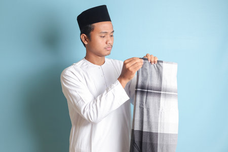 Portrait of Asian muslim man in white koko shirt folding and tidy up his sarung . Isolated image on blue backgroundの写真素材