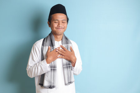 Portrait of religious Asian man in koko shirt or white muslim shirt and black cap, placing hand on heart, feeling very grateful. Isolated image on gray backgroundの写真素材