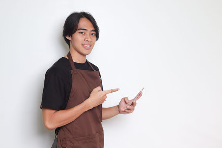 Portrait of attractive Asian barista man in brown apron holding and pointing mobile phone. Isolated image on white backgroundの写真素材