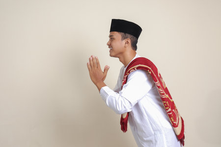 Portrait of attractive Asian muslim man in white shirt with skullcap showing apologize and welcome hand gesture. Isolated image on gray backgroundの写真素材