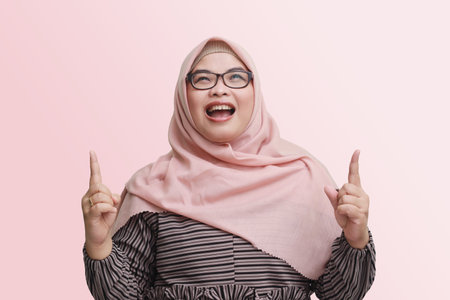 Portrait of cheerful Asian woman with hijab, showing product, pointing at something and smiling. Advertising concept. Isolated image on pink backgroundの写真素材