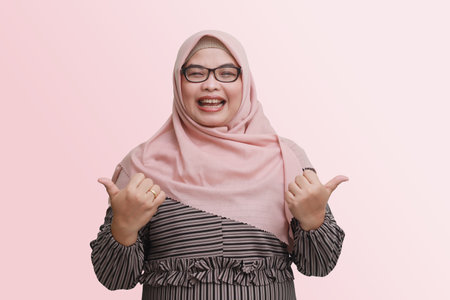 Portrait of cheerful Asian woman with hijab, making thumbs up hand gesture. Isolated image on pink backgroundの写真素材