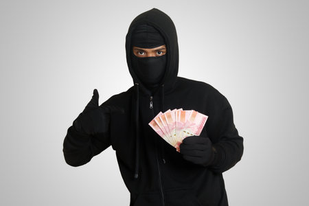 Portrait of mysterious man wearing black hoodie and mask stealing one hundred thousand rupiah successfully from victim. Isolated image on gray backgroundの写真素材