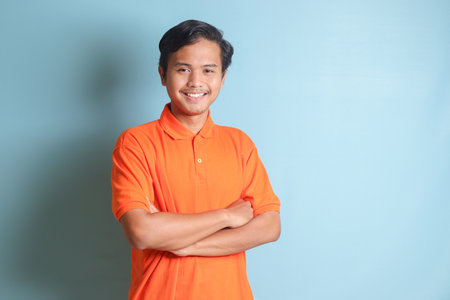 attractive Asian man in orange shirt keeping arms crossed and looking at camera. Isolated image on blue backgroundの写真素材
