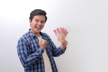 Portrait of excited Asian man in blue plaid shirt standing against white background, showing one hundred thousand rupiah while raising his fist, celebrating success. Financial and savings concept.の写真素材