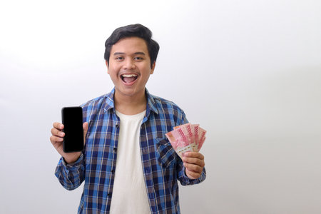 Portrait of excited Asian man in blue plaid shirt standing against white background, showing one hundred thousand rupiah while showing blank screen mobile phone. Financial and shopping concept.の写真素材