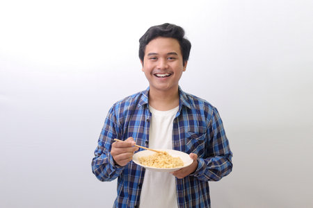 Portrait of happy Asian man in blue plaid shirt eating delicious instant noodles with chopsticks served on plate. Isolated image on white backgroundの写真素材