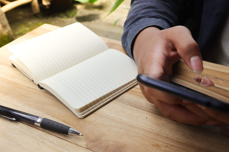 Close up of male hand using mobile phone with empty notebook and a cup of coffee while sitting at...