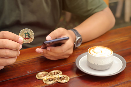 Male hand holding a bitcoin while using phone with a cup of coffee on wooden table inside the cafe. Cryptocurrency trading conceptの写真素材