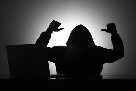 Mysterious man wearing black hoodie using laptop while mocking someone by making thumb down gesture in the dark room. Hacker and cyber security concept.の写真素材