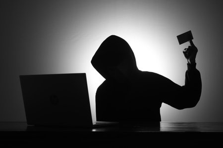 Mysterious man wearing black hoodie using laptop and holding credit card in the dark room. Hacker and cyber security concept.の写真素材