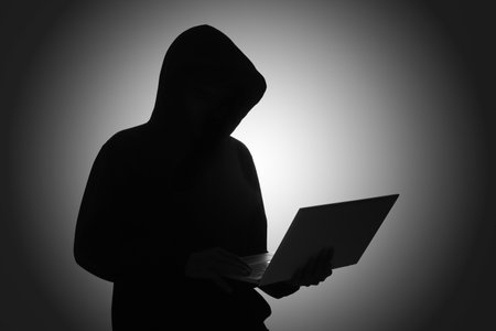 Mysterious man wearing black hoodie using laptop in the dark room. Hacker and cyber security concept.の写真素材