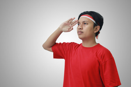 Portrait of attractive Asian man in t-shirt with red and white ribbon on head, giving salute pose...