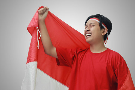 Portrait of attractive Asian man in t-shirt with red white ribbon on head with flag on his shoulder as a cloak, raising his fist, celebrating Indonesia's independence day. Isolated image on grayの写真素材