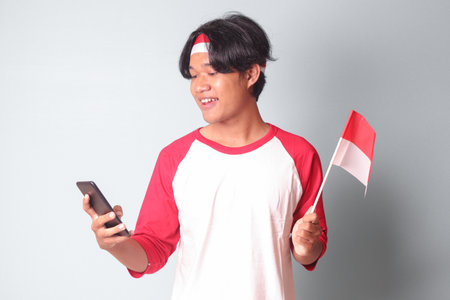 Portrait of attractive Asian man in t-shirt with red and white ribbon on head, holding mobile phone while raising up Indonesia flag. Isolated image on gray backgroundの写真素材