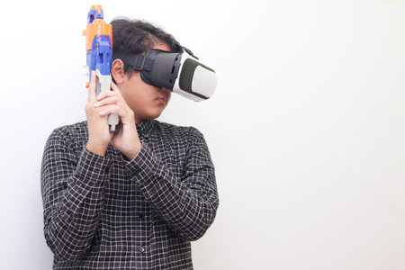 Portrait of Asian man in black plaid shirt using Virtual Reality (VR) glasses holding a gun weapon...