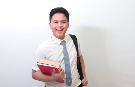 Indonesian senior high school student wearing white shirt uniform with gray tie holding some books, smiling and looking at camera. Isolated image on white backgroundの写真素材