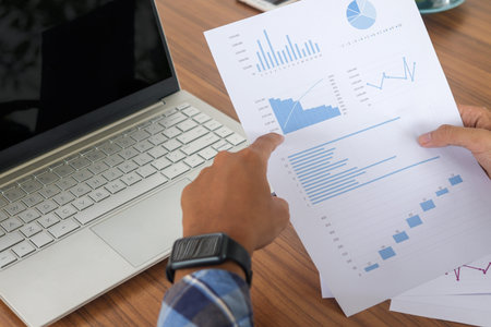 Close up of male freelance worker hand working on laptop and pointing with finger on analysis data with business graph and chart of information diagram on office desk in the morningの写真素材