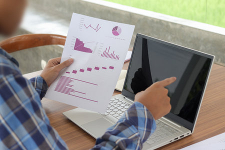 Close up of male freelance worker hand working on laptop and pointing with finger on analysis data with business graph and chart of information diagram on office desk in the morningの写真素材