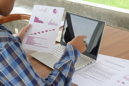 Close up of male freelance worker hand working on laptop and pointing with finger on analysis data with business graph and chart of information diagram on office desk in the morningの写真素材