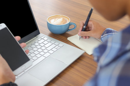 Close up of male hand writing on note book with pen, showing blank screen of laptop and smartphone. Working in cafe concept with a cup of coffeeの写真素材