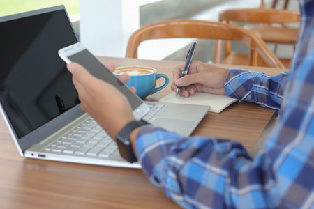 Close up of male hand writing on note book with pen, showing blank screen of laptop and smartphone....