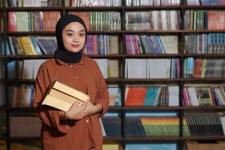 Portrait of Asian hijab woman holding book in front of library bookshelf. Muslim girl reading a book. Concept of literacy and knowledgeの写真素材