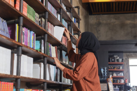 Portrait of Asian hijab woman holding book in front of library bookshelf. Muslim girl reading a book. Concept of literacy and knowledgeの写真素材