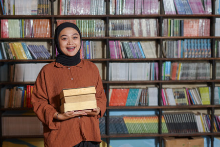 Portrait of Asian hijab woman holding book in front of library bookshelf. Muslim girl reading a book. Concept of literacy and knowledgeの写真素材