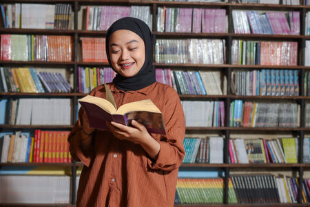 Portrait of Asian hijab woman holding book in front of library bookshelf. Muslim girl reading a book. Concept of literacy and knowledgeの写真素材