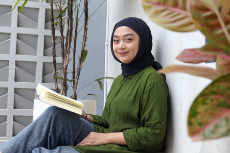 Portrait of attractive Asian hijab woman reading book in balcony. Muslim girl enjoying free time....